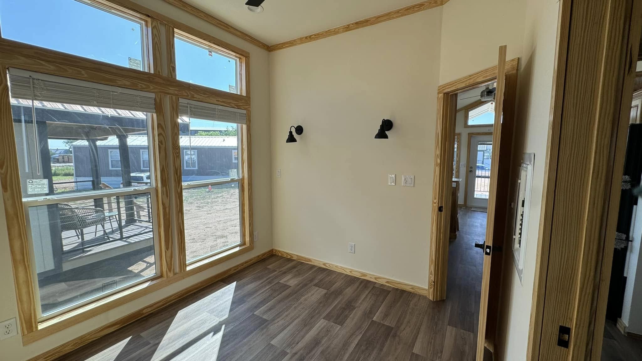Bright room with large windows and wood trim, showcasing a view of outdoor space and neighboring buildings. Walls are off-white with two wall lights.