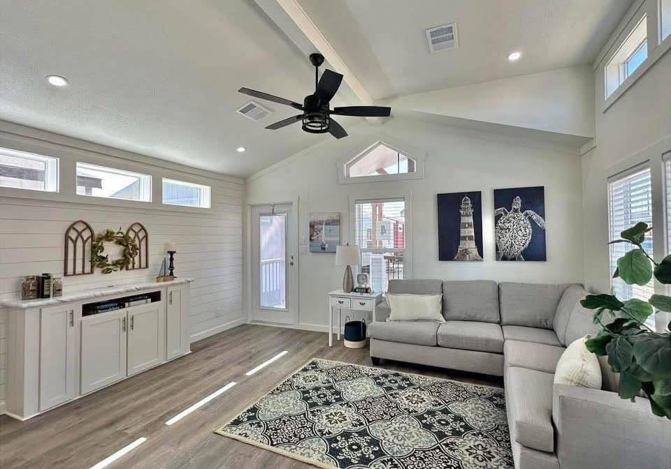 Stylish living room with a gray L-shaped sofa, patterned rug, coastal wall art, and shiplap walls. Natural light filters through numerous windows.