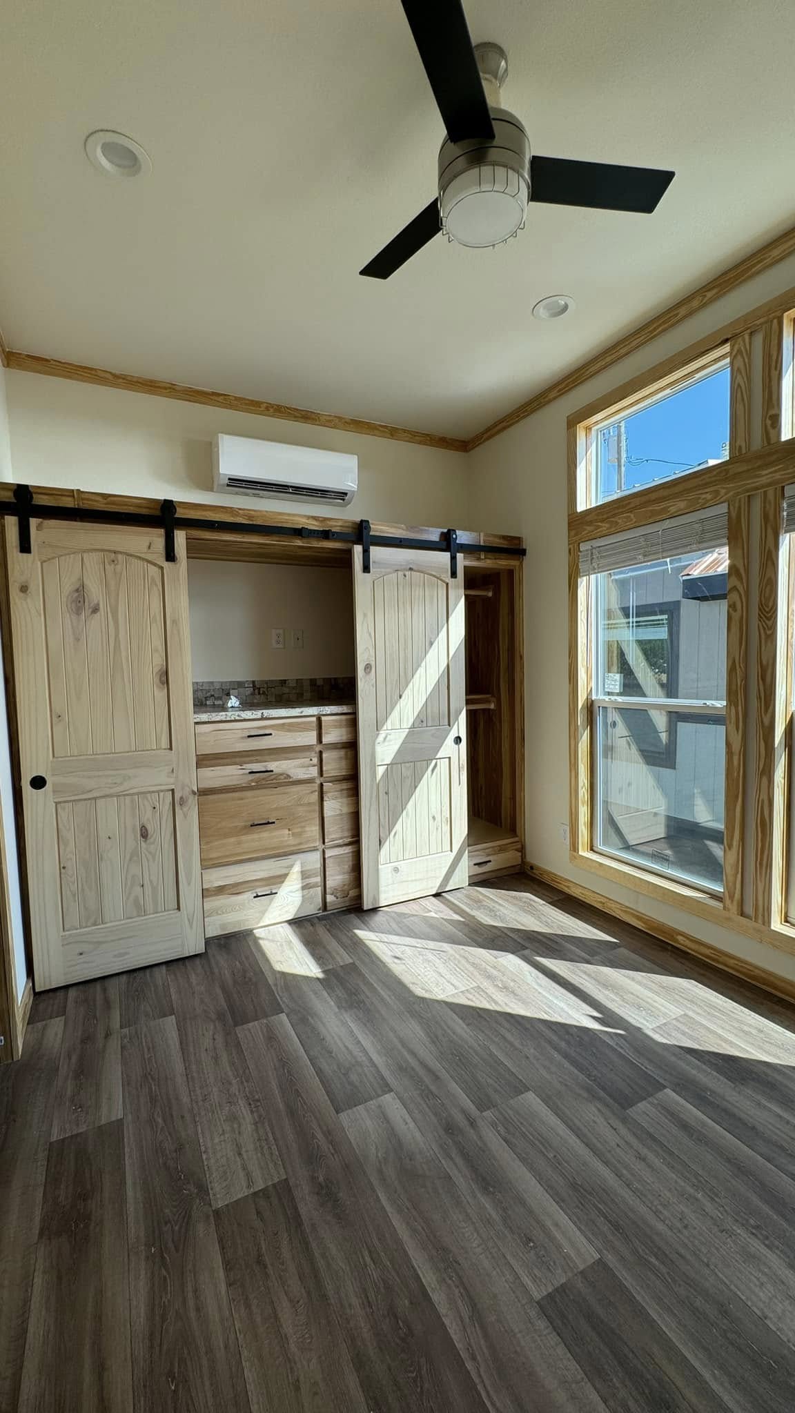 Sunlit room with wooden flooring, featuring sliding barn doors, a ceiling fan, and large windows. The room exudes a bright and cozy ambiance. 