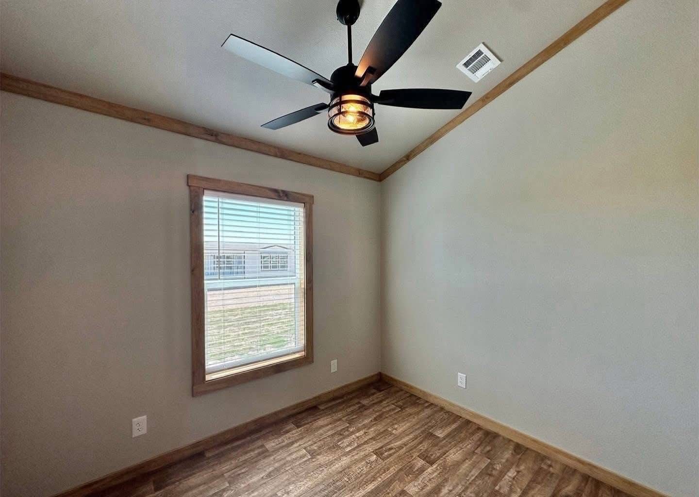 Small empty room with wood flooring, beige walls, and a wooden trim. A large window lets in natural light. A modern black ceiling fan is central.