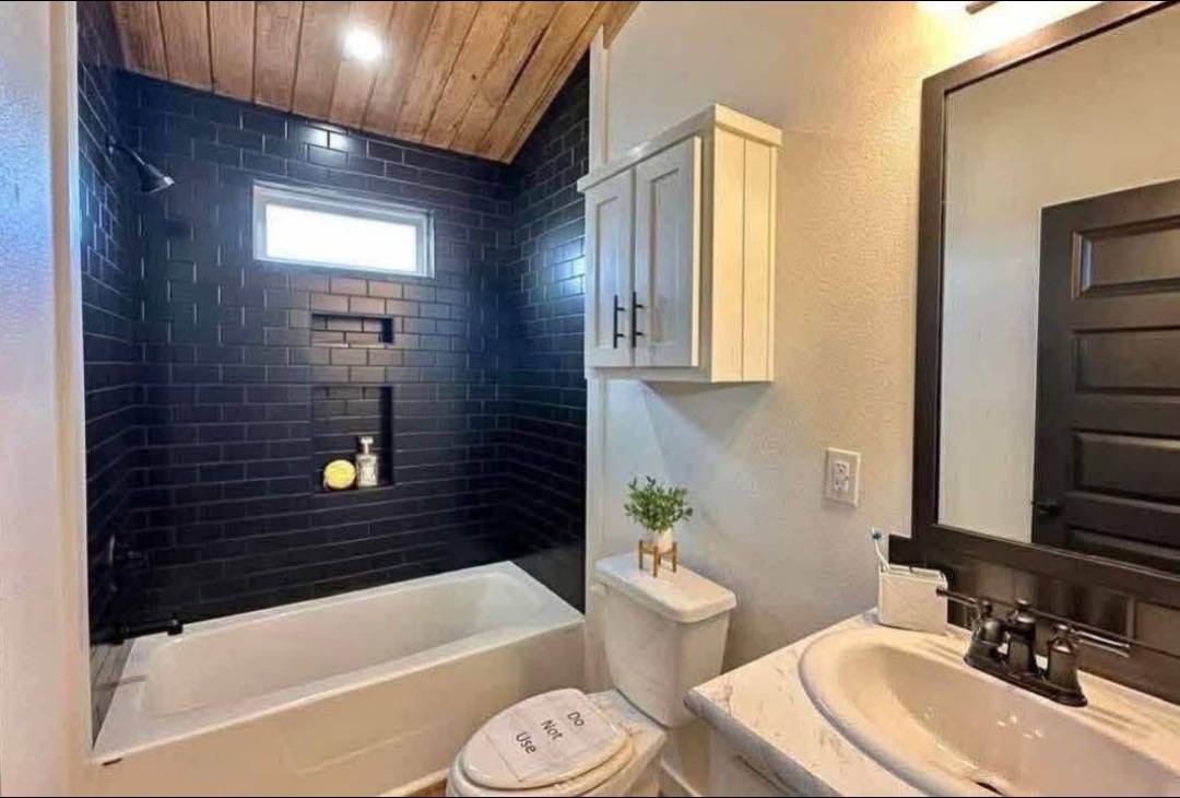 Modern bathroom with a black-tiled shower, small window, white tub, wood ceiling, white vanity, and a sink with a large mirror; cozy ambiance.