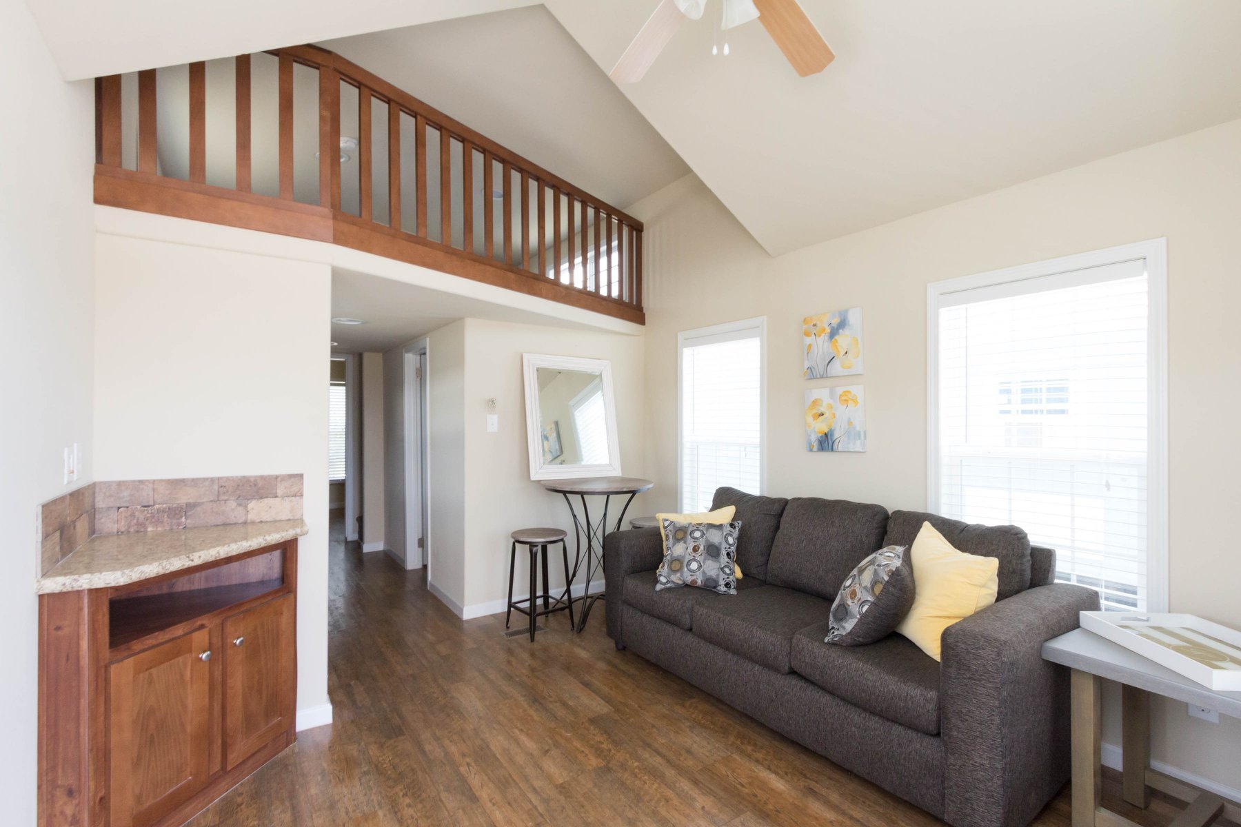 Bright living room with a gray sofa, patterned cushions, and yellow accents. Wood floors, high ceiling, loft railing, and wall art create a cozy feel.