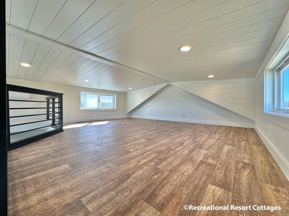 Bright loft space with sloped white ceiling, recessed lights, wood-look flooring, and two windows. A black railing outlines a stairwell.