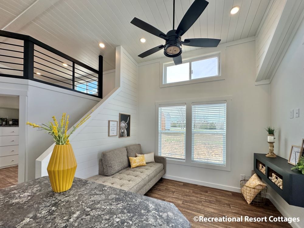 Modern tiny home interior with high ceilings, large windows, gray sofa, and black ceiling fan. A yellow vase with flowers adds a cheerful touch.