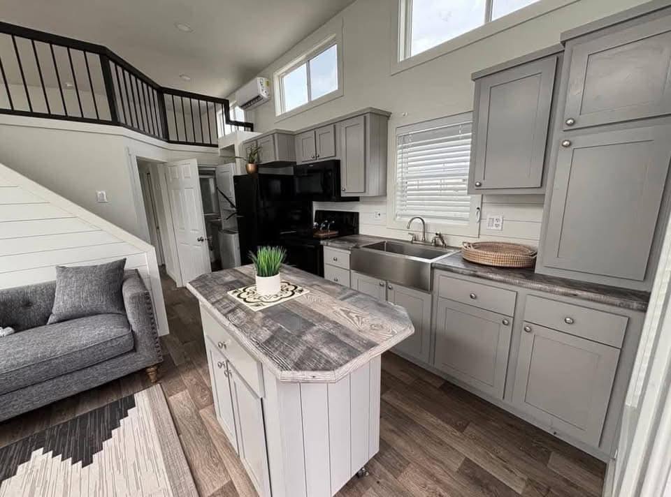 Modern kitchen and living area in a small home. Gray cabinets, rustic island, and a sleek black fridge. Cozy grey couch with stairs leading to a loft.
