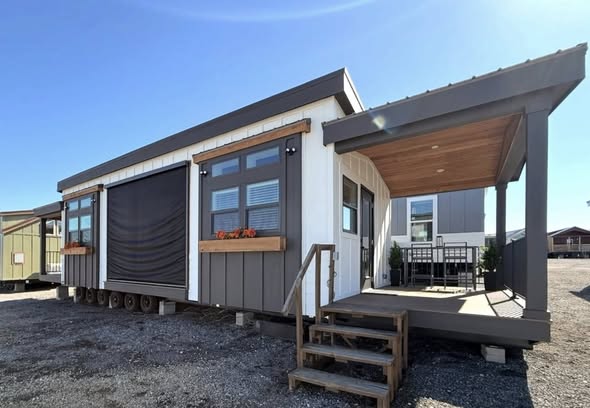 Modern tiny house with a white and gray exterior, wooden accents, and a covered porch with seating. Clear blue sky, creating a serene atmosphere.