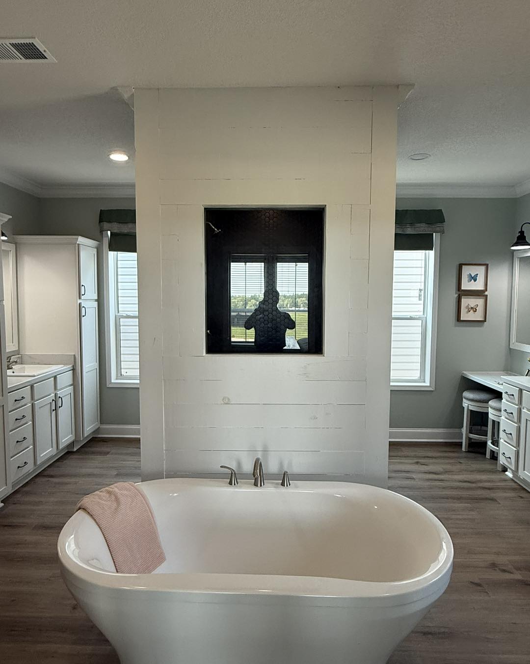 A bathroom with a freestanding white bathtub in the center, a pink towel draped over it. Behind, a mirror reflects a person and two windows. The room appears bright and modern with wooden floors and cabinets.