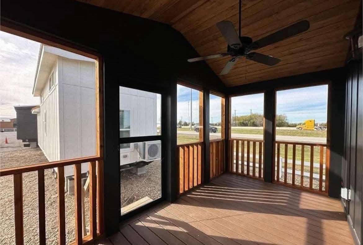 Wooden screened porch with a vaulted ceiling and ceiling fan, overlooking a grassy area and parked trailers. Sunlit with a calm, rustic vibe.