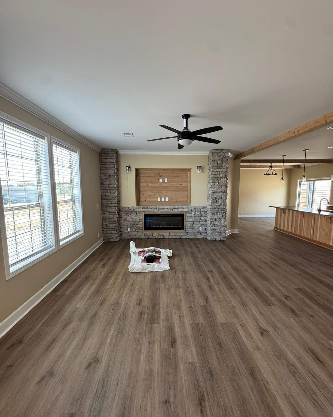 A spacious, empty living room with wood floors, a stone accent wall, and large windows. A ceiling fan hangs above, and construction supplies are on the floor.