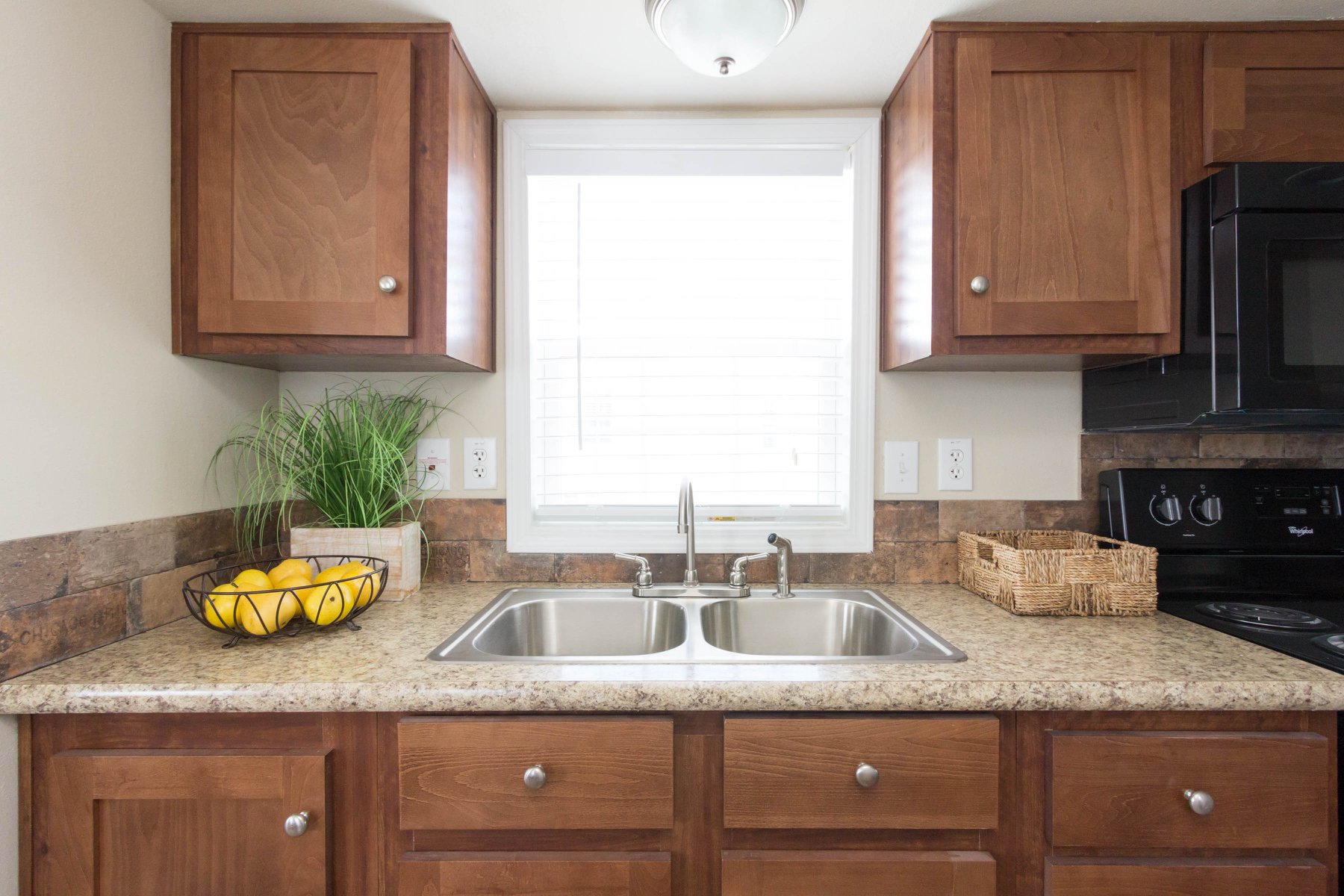 A kitchen with wooden cabinets, a double sink, and a counter. Lemons in a wire basket and green plant next to a window. Cozy and inviting atmosphere.