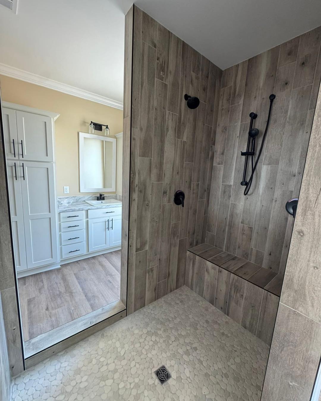 Modern bathroom features a walk-in shower with wood-look tiles and black fixtures, a bench seat, pebble floor, and white cabinetry with a marble countertop.