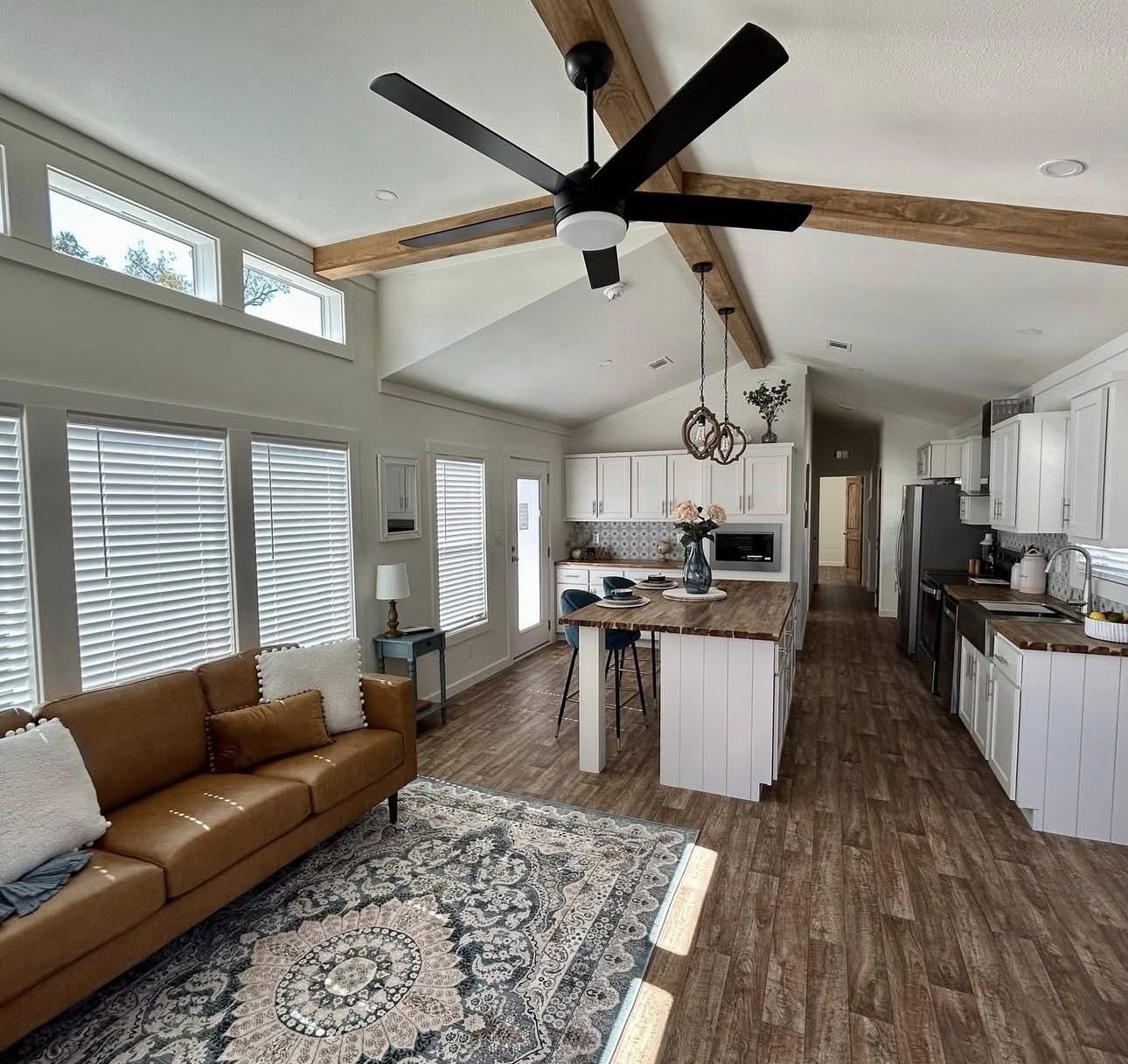 Stylish open-plan kitchen and living room with a high ceiling, wood beams, brown leather sofa, ornate rug, and large ceiling fan, exuding coziness.