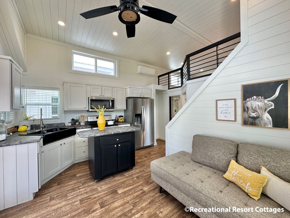 Modern tiny house interior with a cozy living area and kitchen. Features wood flooring, stainless steel appliances, and a loft. Bright, airy feel.