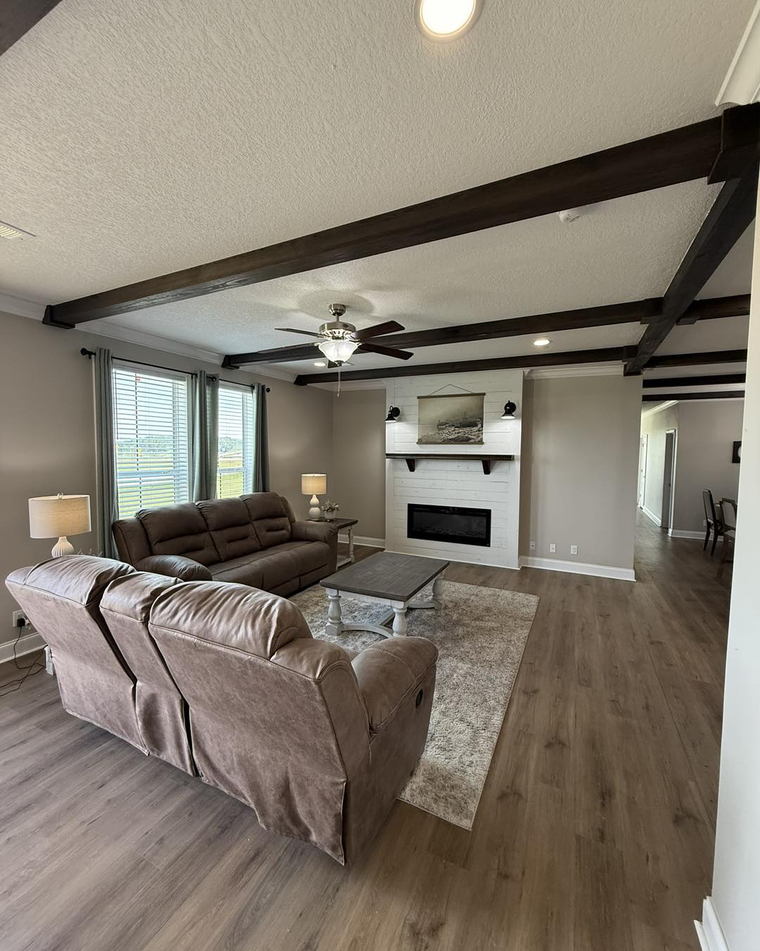 Spacious living room with brown leather sofas, wood beams on the ceiling, and a white fireplace. Natural light floods in through large windows. Calm and cozy atmosphere.