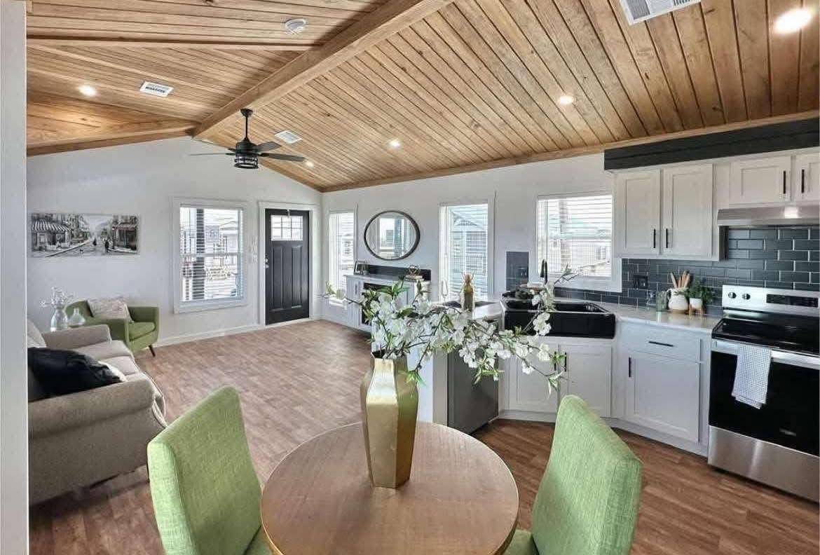 Modern living space with a wooden ceiling, bright kitchen, round table, green chairs, and a beige couch. Warm, inviting, and well-lit atmosphere.