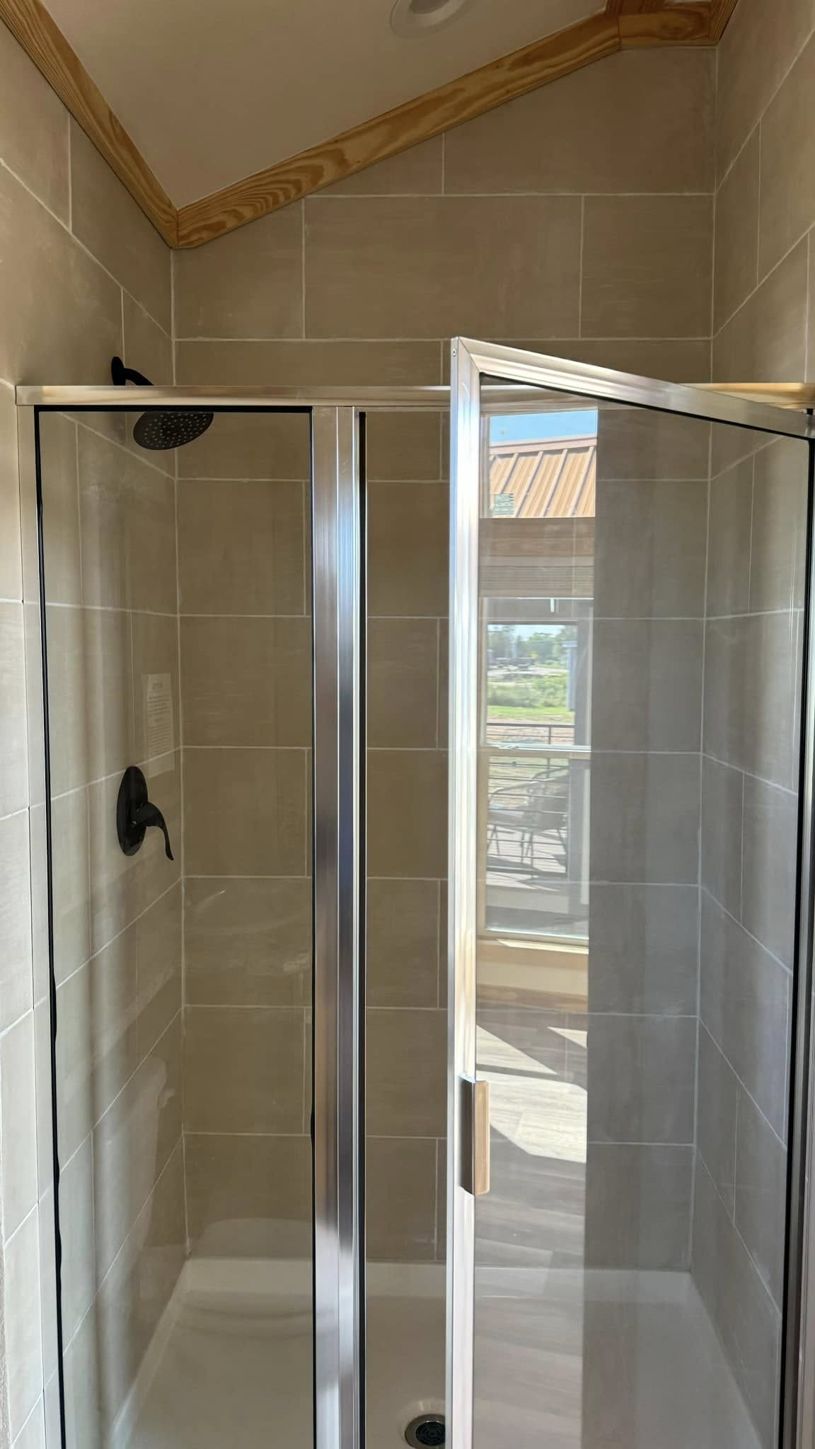 Compact modern shower with beige tiles and chrome fixtures, glass door partially open, wood-paneled ceiling. Sunlight filters through a window.