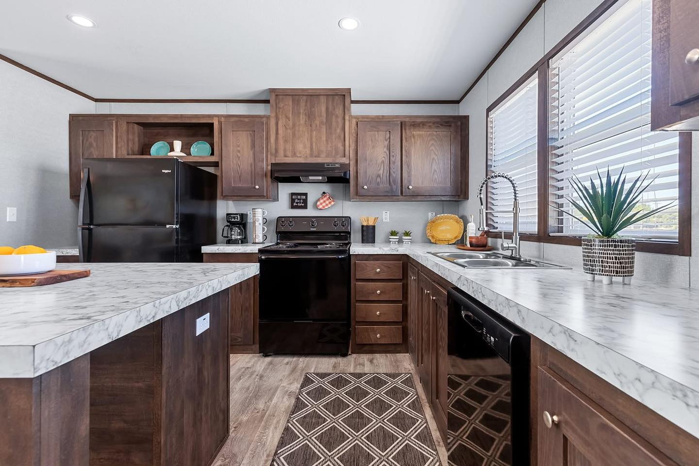 Stylish kitchen with dark wood cabinets, marble-style countertops, black appliances, and decorative items. Sunlight filters through blinds, creating a cozy ambiance.