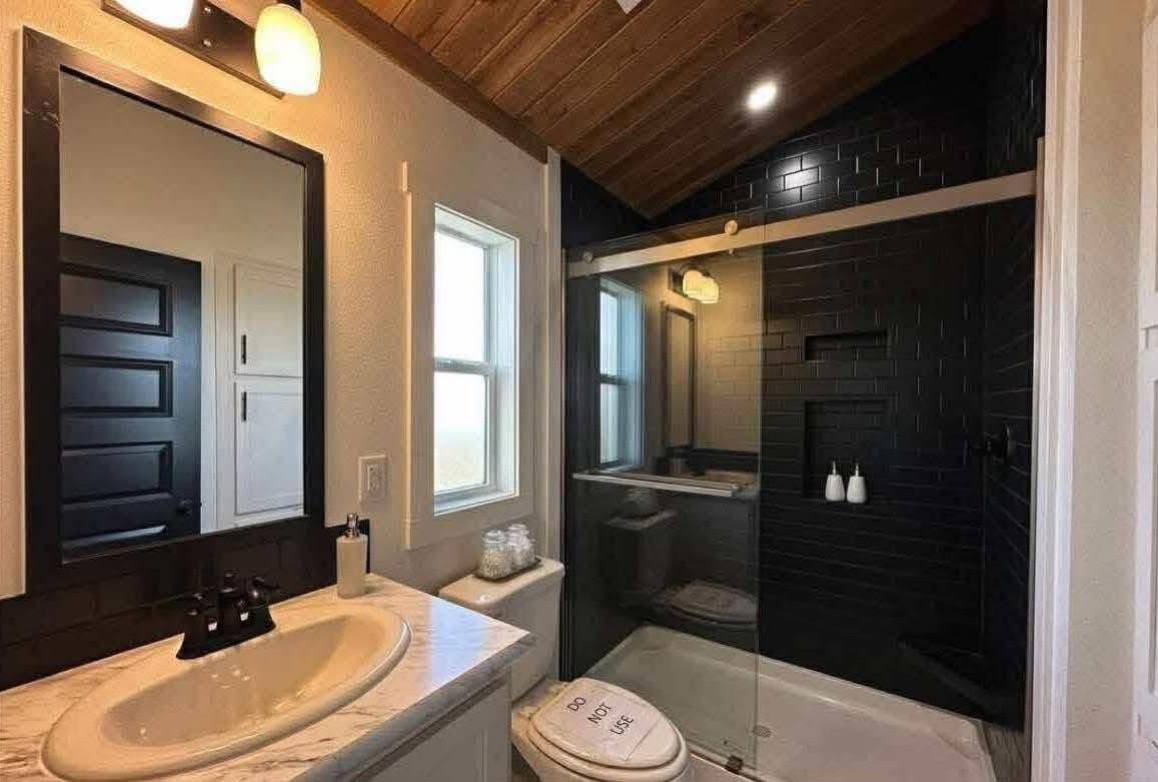 Modern bathroom with wooden ceiling, black-tiled shower, glass doors, a vanity with a white sink, large mirror, and a humorous toilet cover reading "Do Not Look."