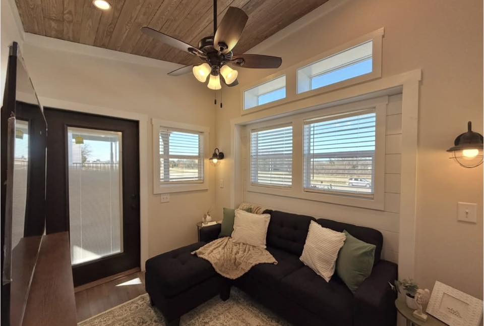 Cozy living room with a dark, L-shaped sofa adorned with pillows and a throw. Large windows let in natural light. Ceiling fan and rustic decor create a warm atmosphere.