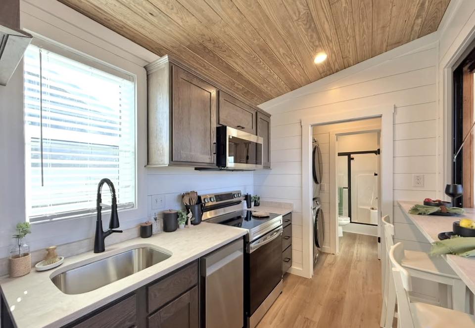 Cozy kitchen with wood ceiling, white walls, and light wood flooring. Dark wood cabinets, stainless steel appliances, and a large window create a warm, inviting atmosphere.