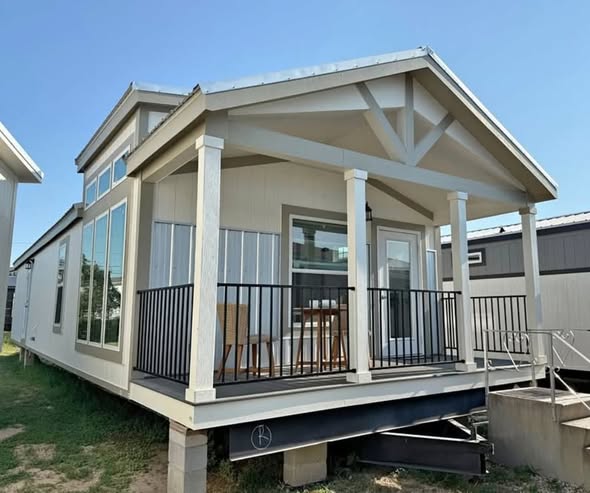 A small, modern tiny house on stilts with a gabled roof and large windows. The porch has a black railing, table, and chairs, evoking a cozy atmosphere.