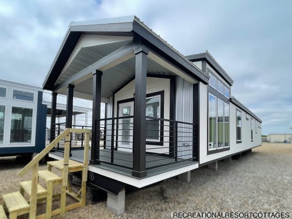 Modern tiny home with a black and white exterior, featuring a small porch with wooden steps and large windows. Positioned on a gravel lot under a cloudy sky.