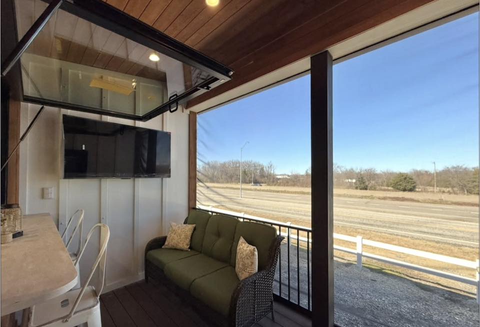 Outdoor patio with a green couch, white pillows, and a wall-mounted TV. Open view of a road and clear sky, conveying a relaxed, inviting atmosphere.