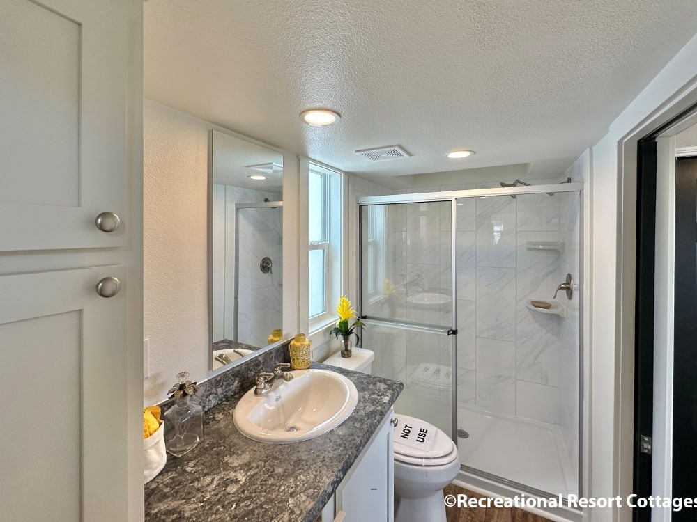 Modern bathroom with a marble countertop and sink, glass-enclosed shower, white cabinets, and bright lighting. A yellow flower adds a cheerful touch.
