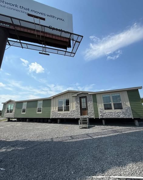 A large green and cream manufactured home with a stone accent exterior sits on a gravel lot under a blue sky. A billboard is visible above.