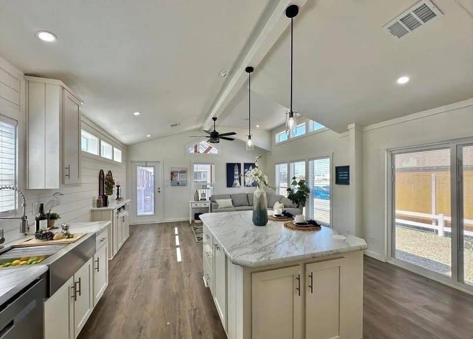 Modern kitchen and living area with white cabinets, marble countertop island, hardwood floors, and large windows, conveying a bright and airy atmosphere.