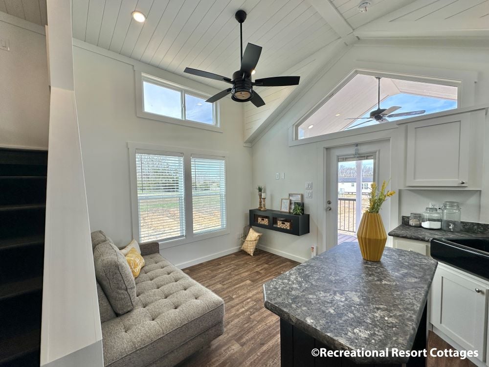 Bright, cozy living room with high ceilings, a ceiling fan, large windows, gray sofa, modern decor, and open kitchen. Sunlight and a calm ambiance.