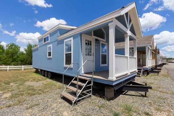 A light blue tiny house on wheels with white trim, three steps leading to a small porch. It’s set on a gravel path under a bright blue sky.