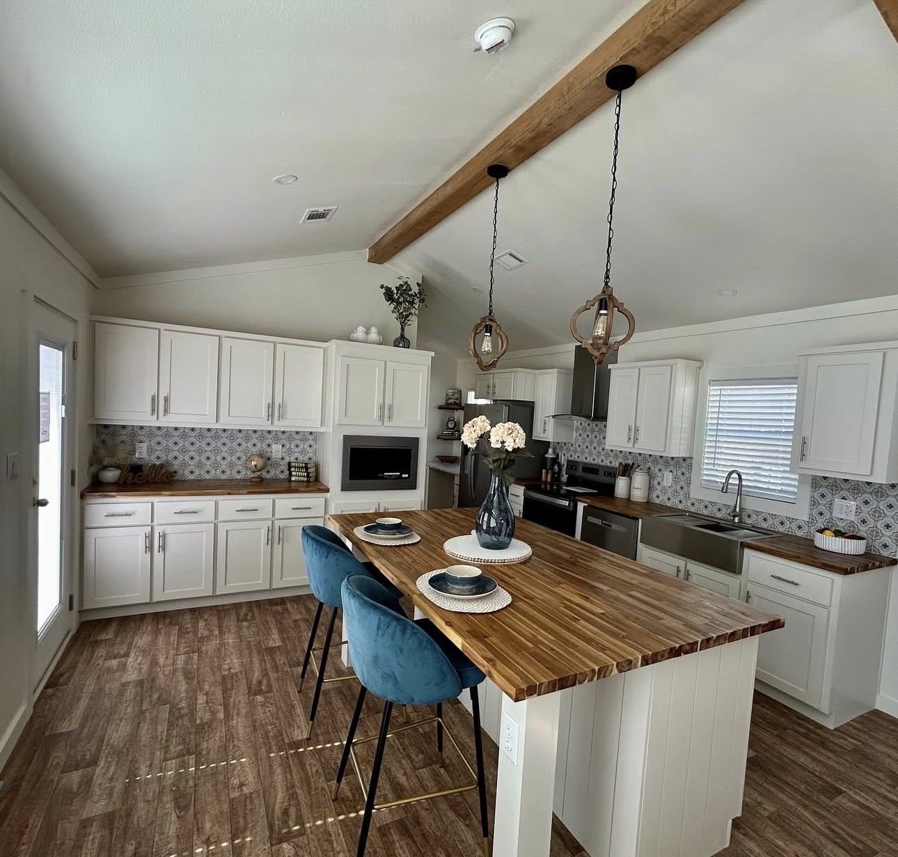 Modern kitchen with wooden accents and white cabinets. A wooden island has blue chairs and a vase of flowers, under two pendant lights. Cozy, inviting space.