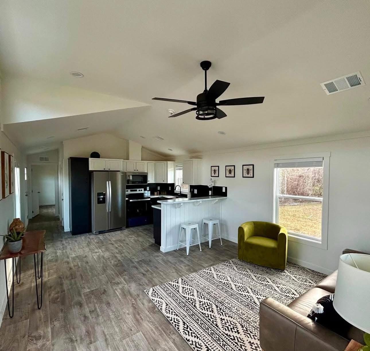 Modern open-plan living room and kitchen with wooden floors, white walls, and a ceiling fan. Features a cozy green chair, patterned rug, and large window.
