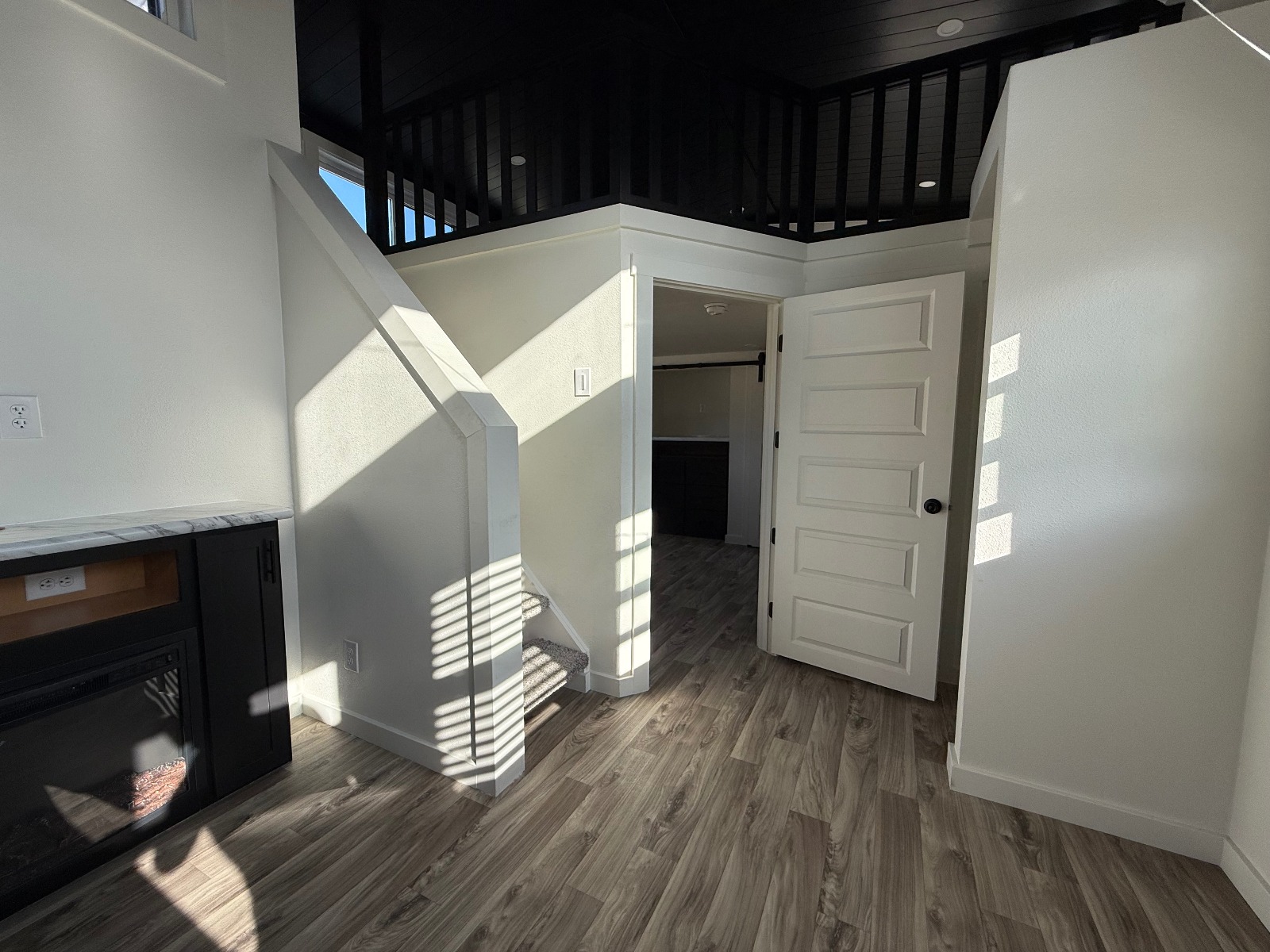 A small, modern living space with light wood flooring, white walls, a black fireplace, and an open door leading to a loft with a dark railing. Natural light casts shadows, creating a cozy atmosphere.