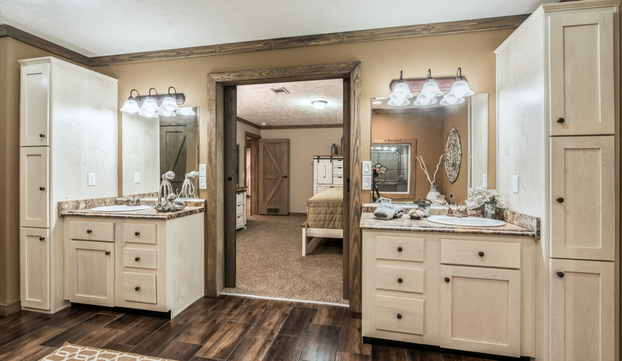 Spacious bathroom with dual vanities, light wood cabinets, and granite countertops. Warm brown tones, dim lighting, and a view into a cozy bedroom.