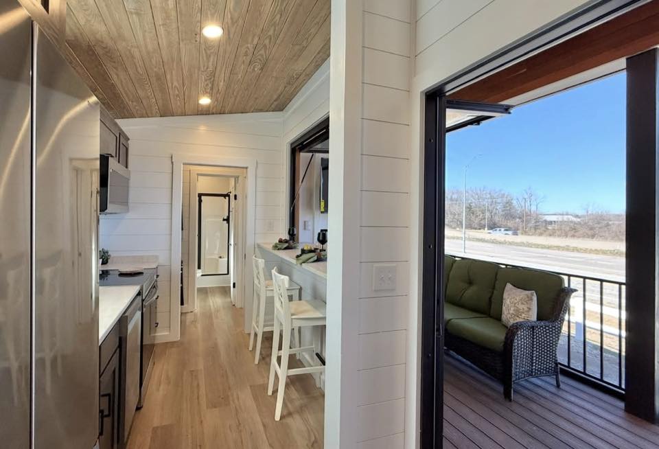 Narrow modern kitchen with wood ceiling and light floors, leading to a small porch with green outdoor furniture. Bright and cozy atmosphere.