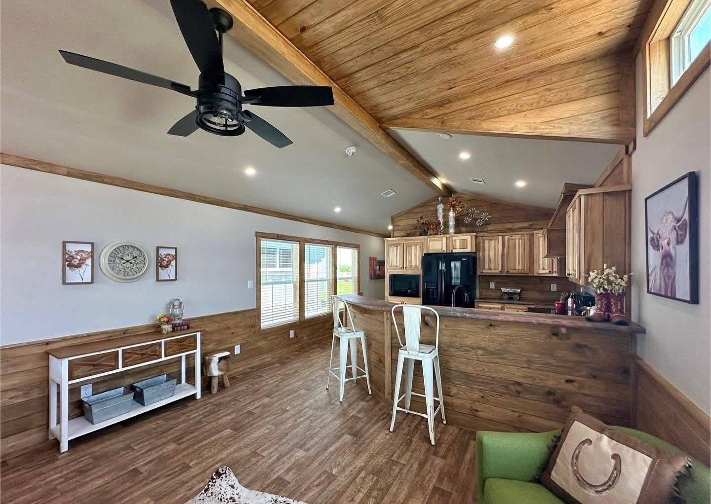Spacious living area with wooden floors and ceiling; features a ceiling fan, bar with stools, floral decor, and a wall clock, creating a rustic vibe.
