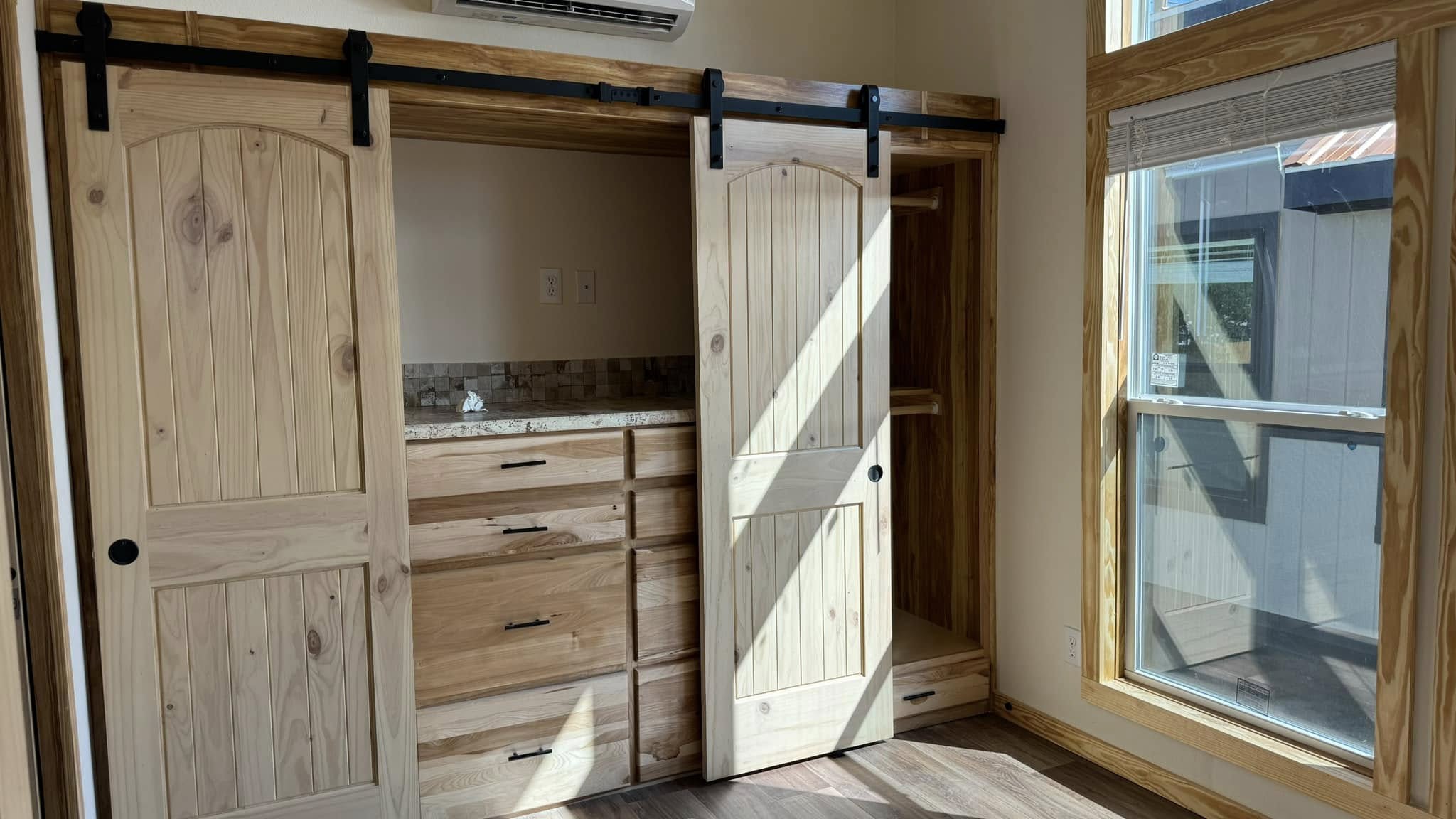 Rustic room featuring wooden sliding barn doors revealing a closet with drawers. Sunlight streams through a large window, creating a warm, inviting ambiance.