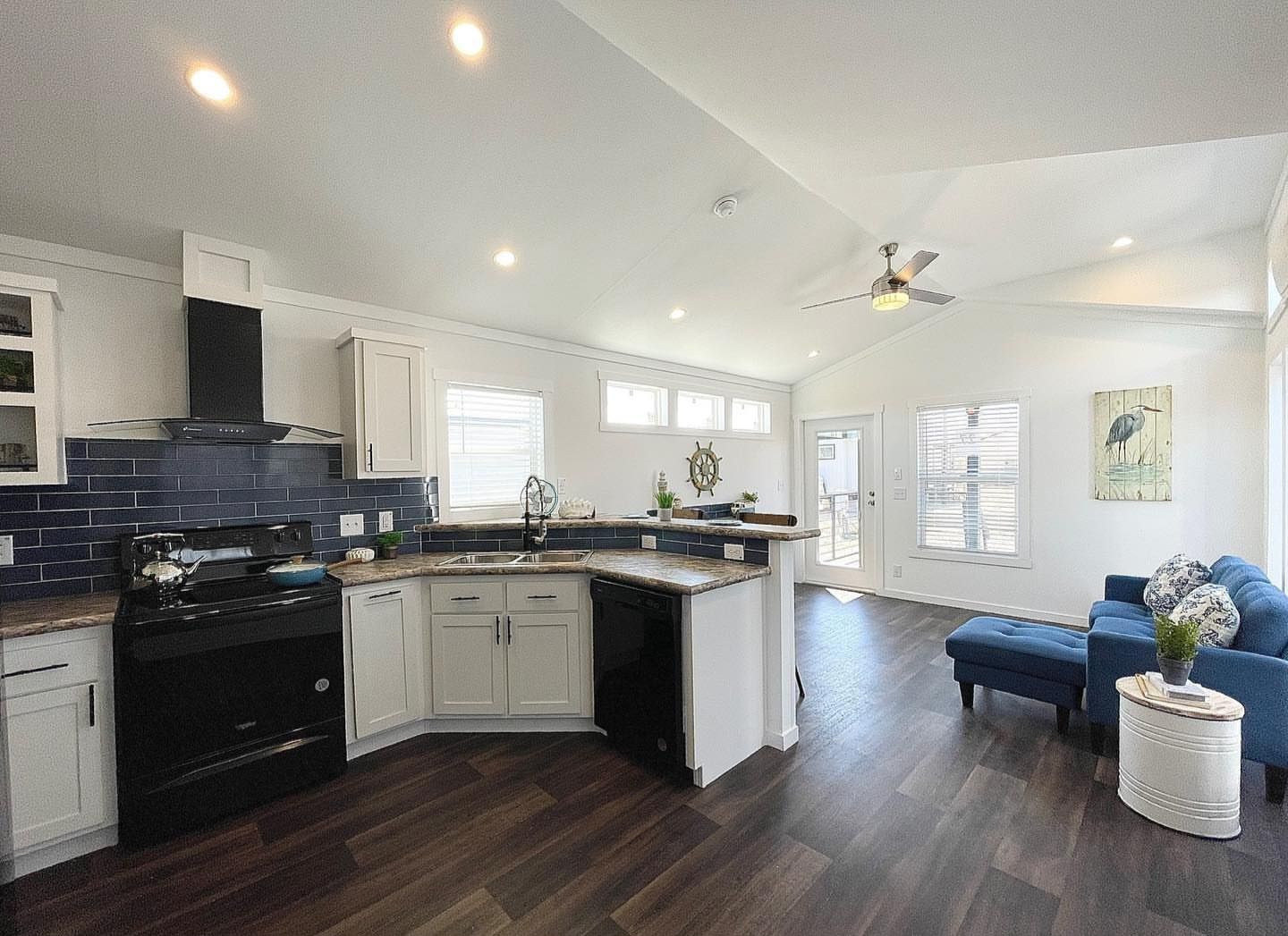 Modern open-concept living room and kitchen with high ceilings. Features dark wood floors, navy backsplash, white cabinets, and blue sofa set. Bright and airy.