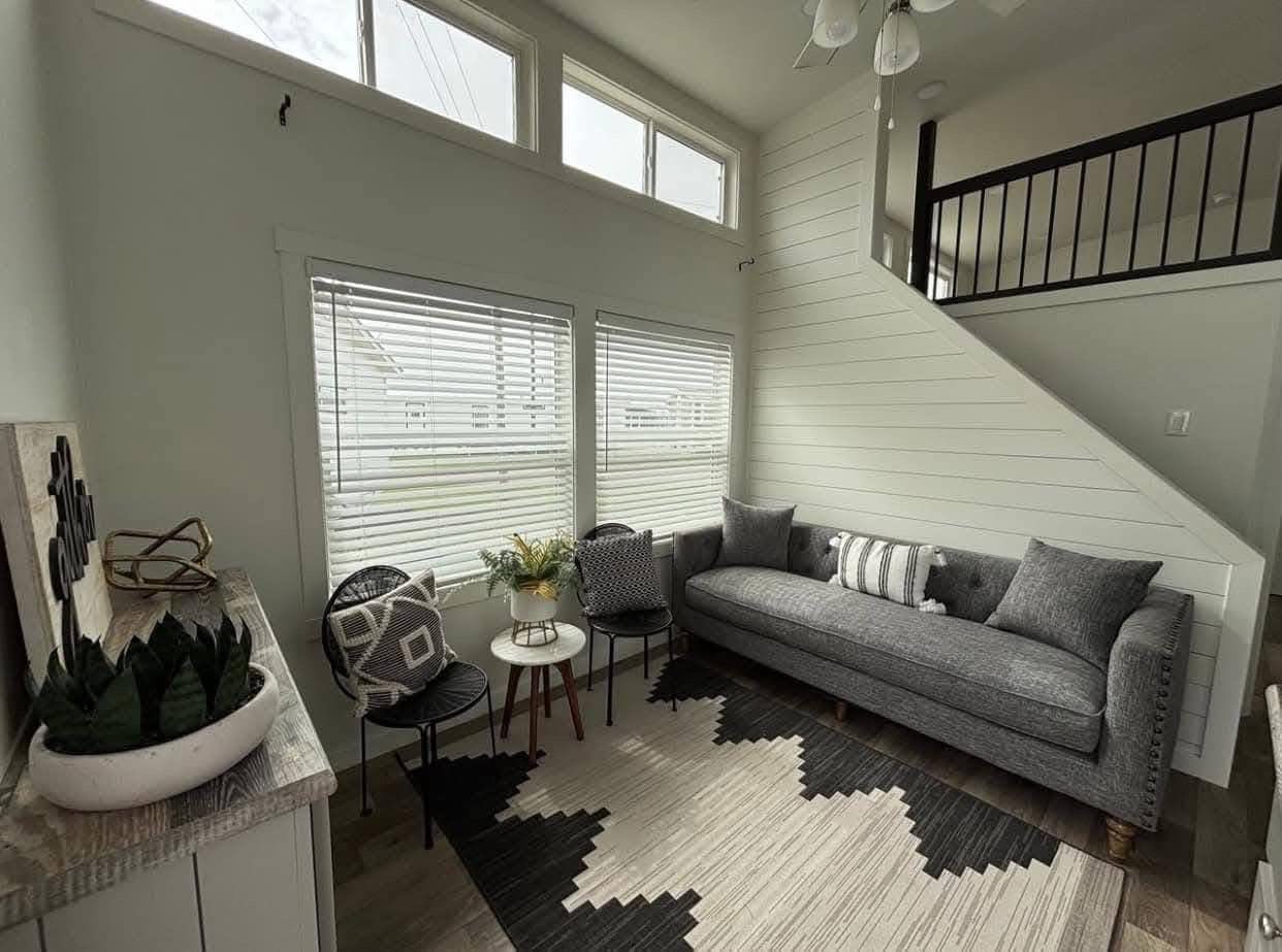 A cozy living room with a modern gray couch, patterned cushions, and a geometric rug. Light streams through large windows, creating a bright, inviting space.