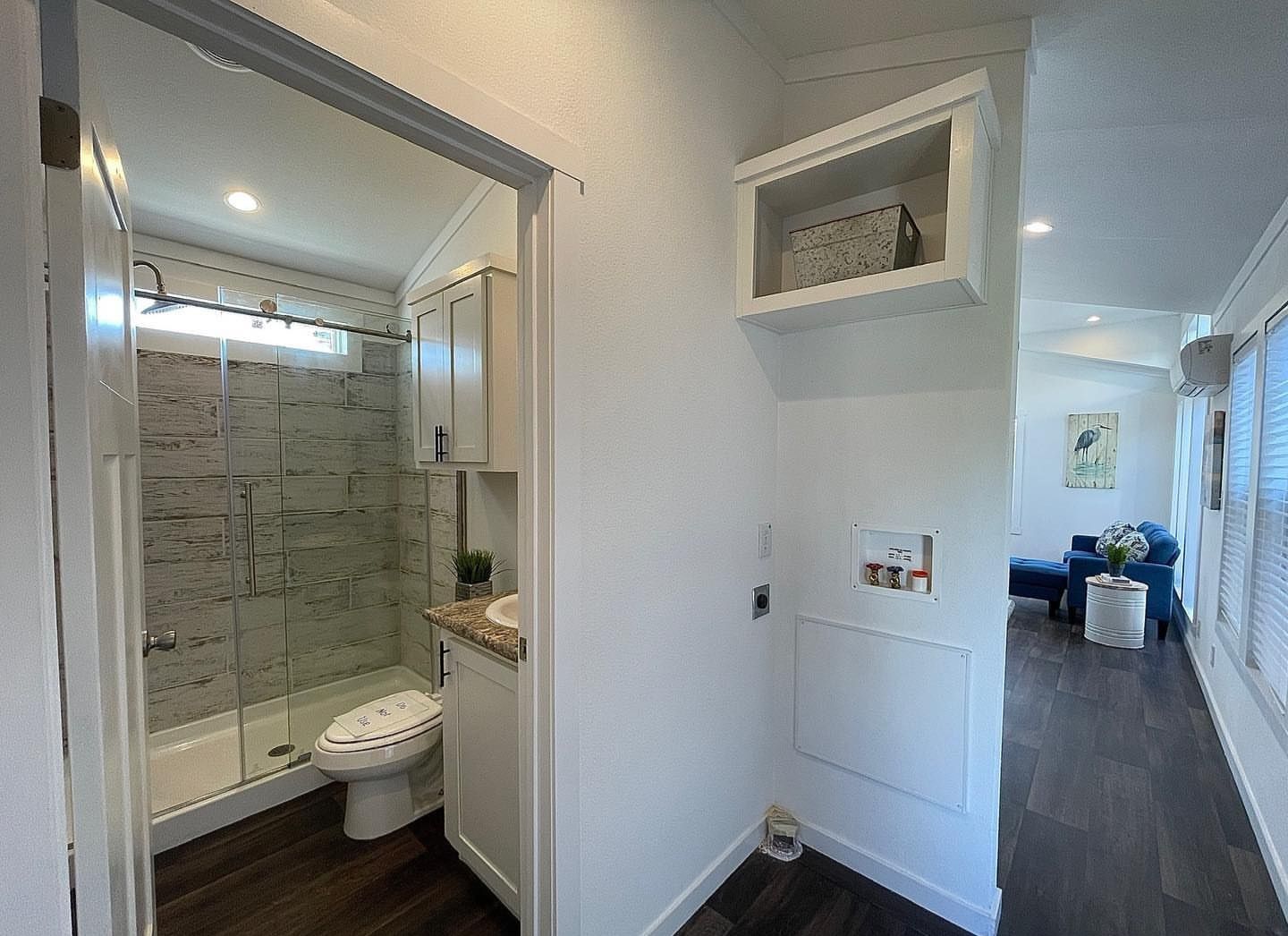 Bright, modern bathroom with stone-textured shower tiles and wood floor, adjacent to a hallway leading to a cozy living area with a blue sofa.