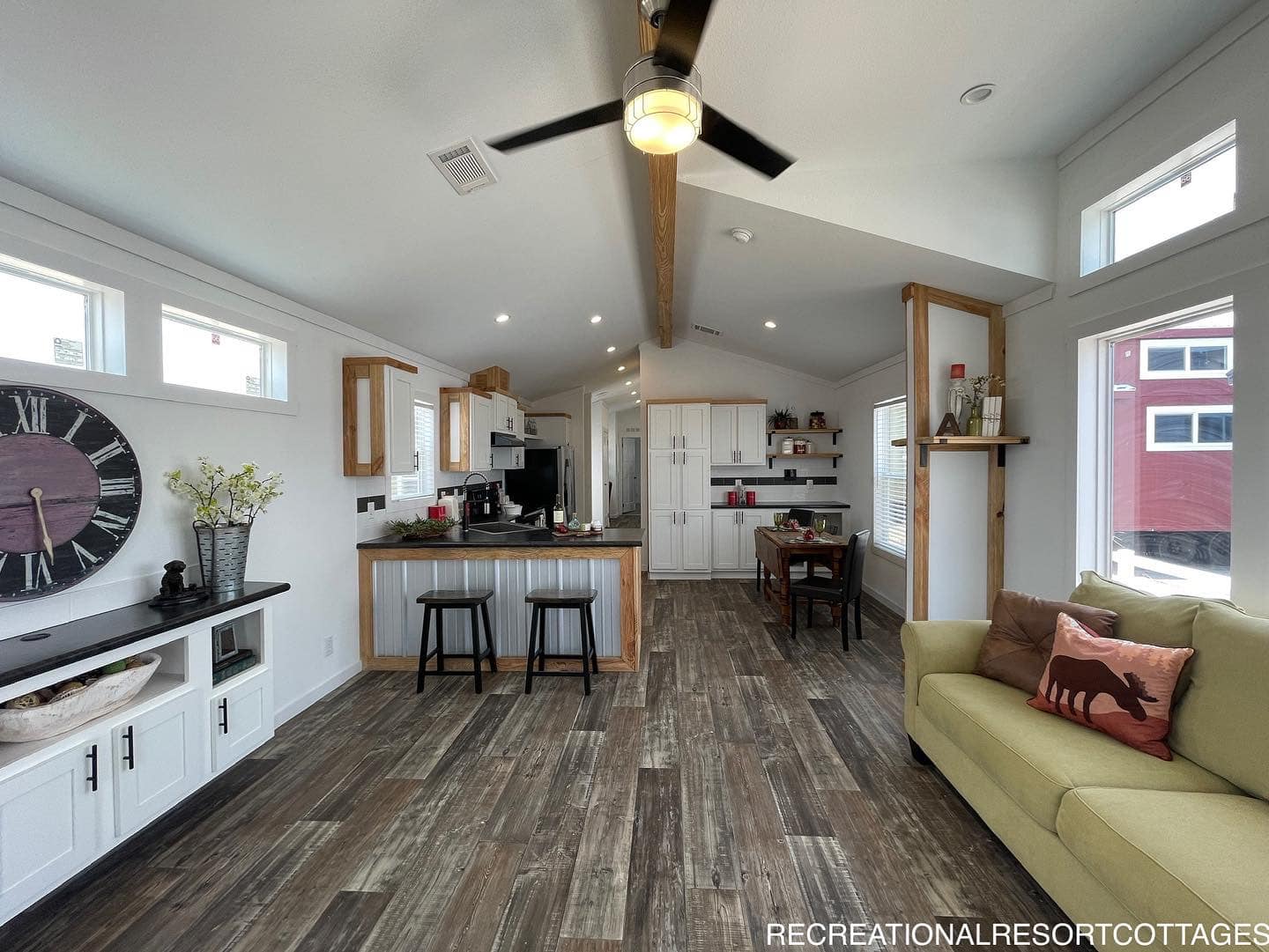 Spacious living area with wood flooring, light walls, and natural light from large windows. Features a kitchen island with stools, a dining area, and ceiling fan. Cozy, modern ambiance.