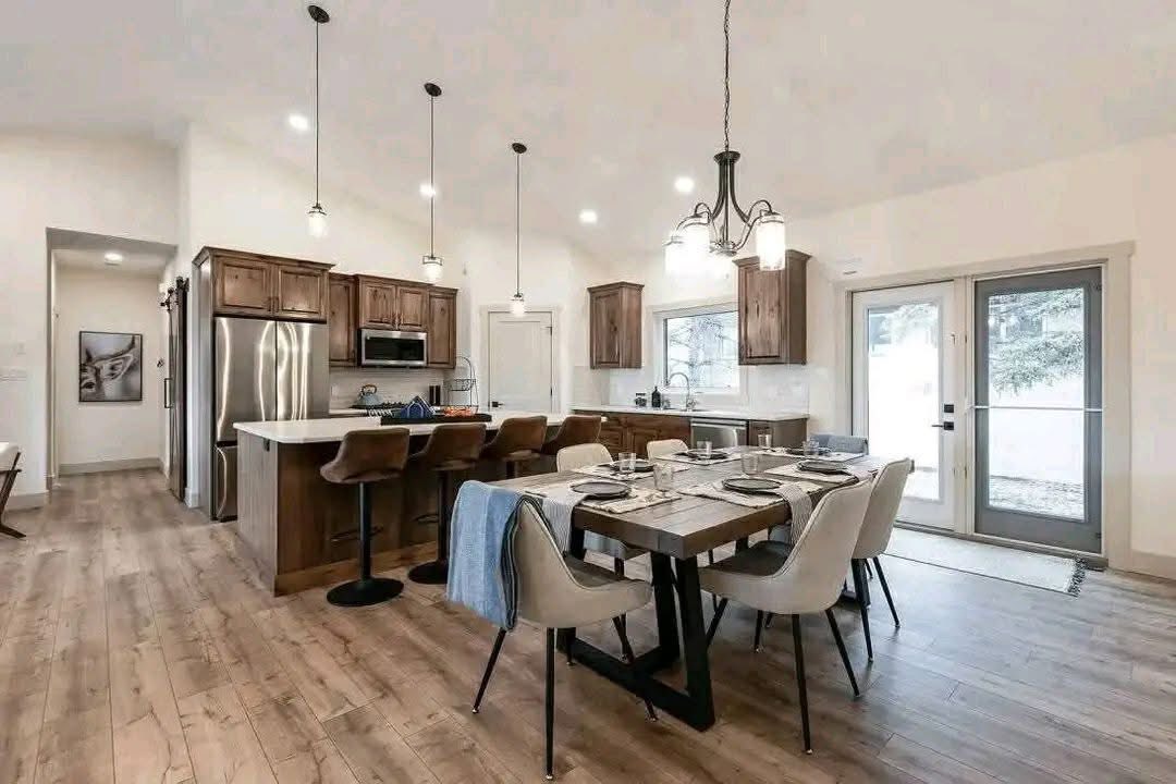 Spacious kitchen and dining area with wooden flooring, featuring a modern table set for six. Pendant lights and large windows convey a cozy, inviting atmosphere.