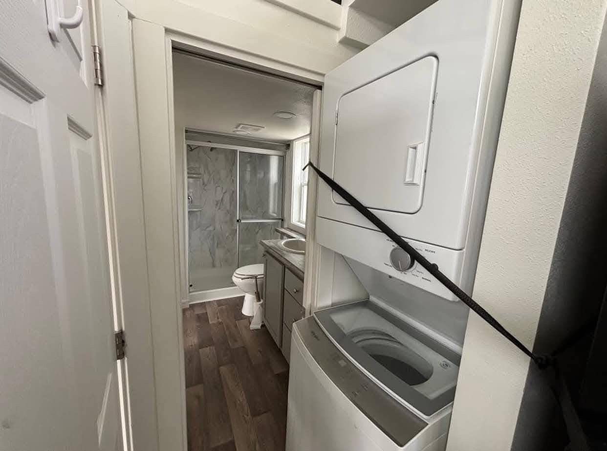 Compact bathroom and laundry area with white stacked washer and dryer on the right, glass-enclosed shower, sink, and toilet on wood flooring.