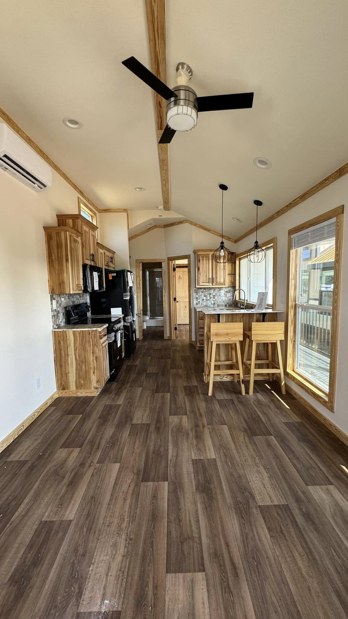 Modern kitchen with wood floors, light wooden cabinets, and a breakfast bar with stools. Ceiling fan above, large window on the right, bright and inviting.