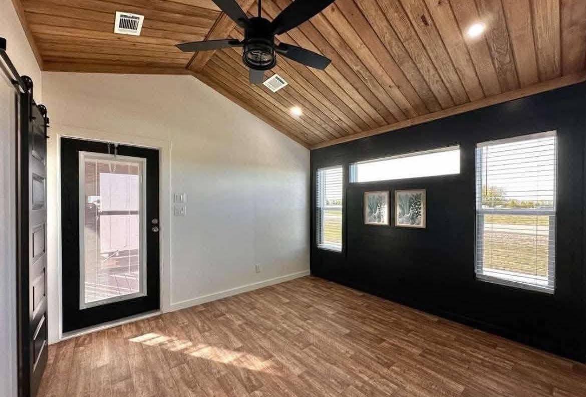 Modern room with a wooden ceiling and floor, black accent wall, large windows, and ceiling fan. Bright natural light enhances the minimalist decor.