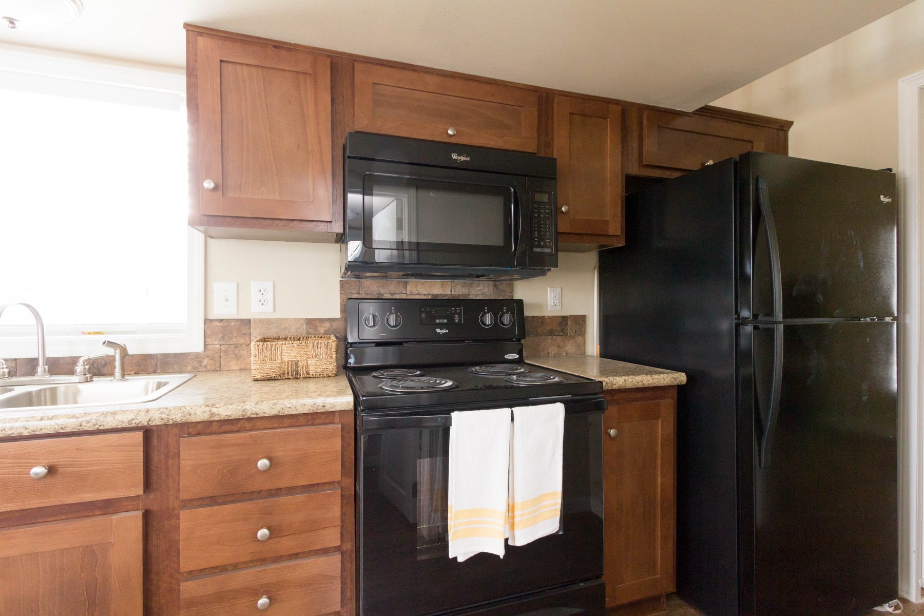 Compact kitchen with wooden cabinets, black appliances including a microwave and refrigerator, a beige countertop, and a stainless steel sink.