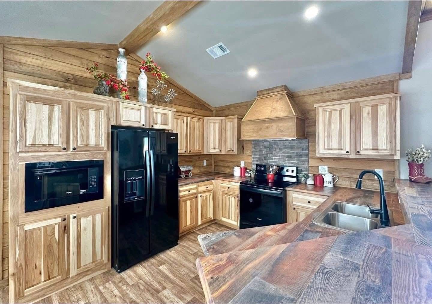 Rustic kitchen with wooden cabinets, black appliances, and a dark tiled backsplash. Decor includes ceramic vases and red accents, creating a cozy vibe.
