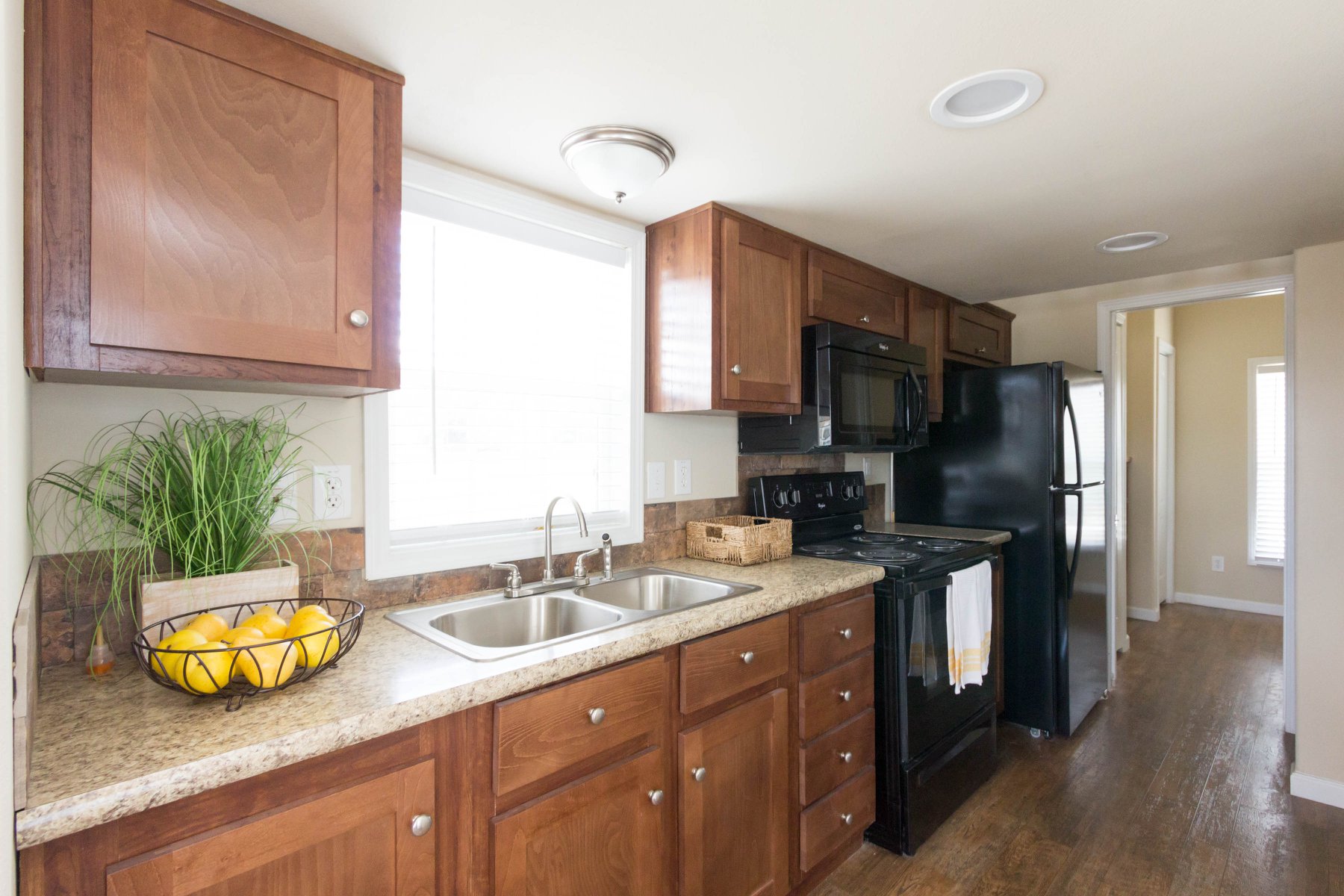 Compact kitchen with wooden cabinets, granite countertops, and stainless steel sink. A bowl of lemons and a plant add color. Black appliances create contrast.